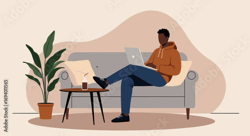 Young man relaxing on a sofa at home, working remotely on his laptop with a drink nearby. Depicts modern work-life balance and comfort.