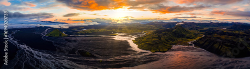 Billede på lærred Aerial view of a green volcanic crater, dark sands, braided glacial rivers, mossy hills, and distant glaciers under a vibrant sunset sky in Iceland