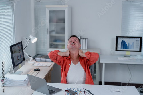 Office worker experiencing neck pain while sitting at desk with laptop, computer and paperwork, demonstrating the physical strain and discomfort associated with prolonged desk work