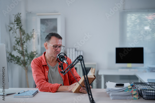 Photos Middle aged male podcaster reading book into microphone while recording podcast