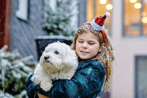 Little school girl playing with little maltese puppy outdoors in winter. Happy child and family dog having fun with snow. Cold weather, snowing.