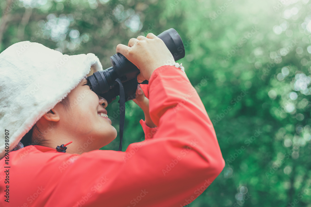 Fototapeta premium A smiling girl wearing red shirt and white hat holds a binoculars looks out the green forest