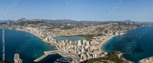 The Panoramic Coastline of Calpe, Spain. Sandy beaches stretch along the clear, turquoise waters on either side, while mountains and hills form a scenic backdrop.