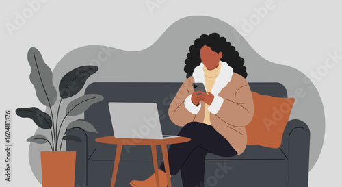 Woman in a cozy coat uses her smartphone while relaxing on a sofa with a laptop nearby, depicting remote work or casual online activity at home.