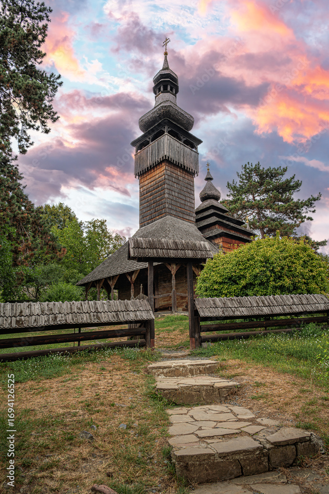 Fototapeta premium A traditional wooden church with a tall shingled tower stands surrounded by trees and a rustic fence under a colorful sunset sky, creating a serene and picturesque rural scene.