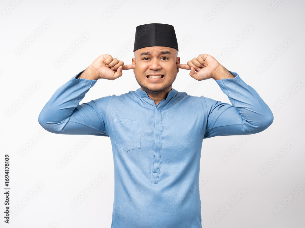 Fototapeta premium Angry boring young asian muslim man headache stressed and frustrated covering his ears with hands due to loud noise. Annoyed noise sound pollution blocking ears. Islam wearing songkok muslim costume