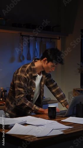 Young man working late night on laptop in a dimly lit kitchen, focusing on tasks with papers