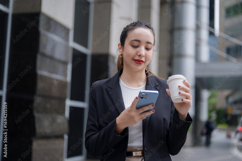Fototapeta premium A smiling office woman is standing outside an office building, holding a cup of coffee while checking her smartphone.