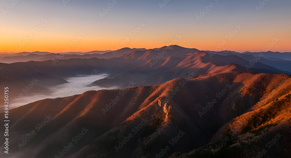 Fototapeta premium A breathtaking panoramic view of a majestic mountain range during a serene golden hour sunrise, with warm light illuminating the peaks and a sea of fog resting in the valley below