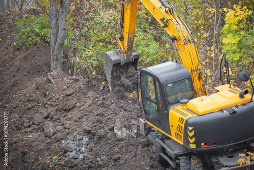 Excavator digs soil with bucket at construction site with trees under difficult working conditions.