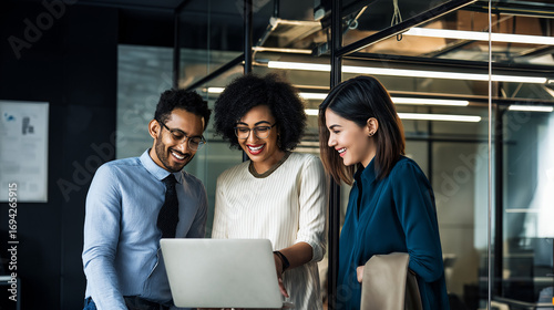 three of diverse colleagues collaborating on laptop near office framework structure, discussing ideas and smiling, modern workplace environment with glass and metal elements, teamwork and innovation c