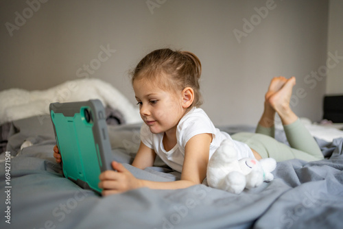Little girl lying on her parents bed and watching cartoons on a tablet. Concept of screen time, digital entertainment and relaxation during early childhood.