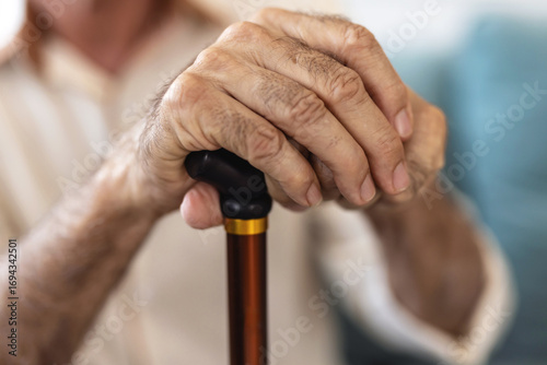 Senior man sitting on the sofa and looking contemplative while holding his walking stick. Walking difficulties. Serious bearded senior leaning on walking stick while thinking about his helplessness.
