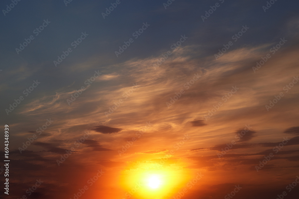 Fototapeta premium Beautiful colorful dramatic sky with clouds at sunset. Bright orange setting sun low on the horizon. Cirrus clouds change color from blue to yellow to red. Stunning backdrop, gorgeous scenery.