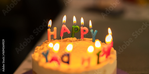 Close-up of a birthday cake decorated with colorful candles spelling the word happy, glowing with warm light in a dark background. Concept of celebration, party, happiness, and special moments