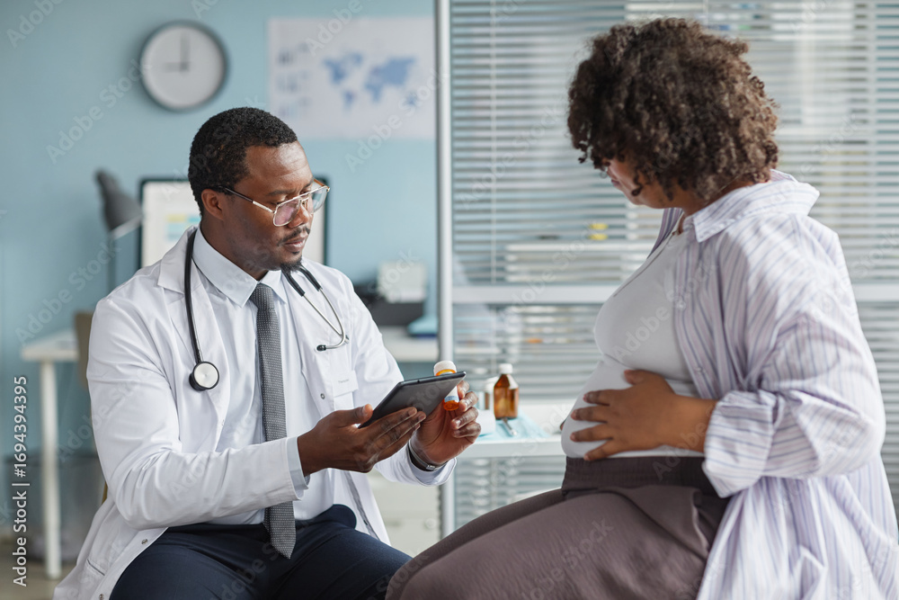 Obraz premium Black middle aged male doctor holding digital tablet consulting pregnant Black woman sitting in medical office, both focused on discussion, modern healthcare setting visible in background