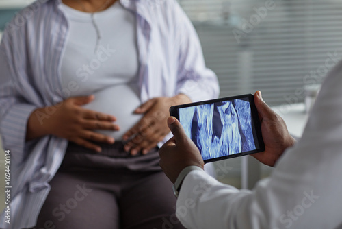Pregnant Black woman sitting with hands on belly while medical professional holding tablet displaying ultrasound image during prenatal checkup in clinical setting