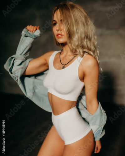 Elegant young woman with long blonde hair in white sportswear and denim jacket posing indoors, looking confident and stylish in soft studio light.