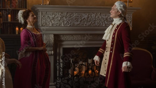 Medium full shot of polite Victorian era male aristocrat, dressed in red surcoat, breeches and wig arriving in manor to visit two lady friends, greeting, bowing and kissing hands