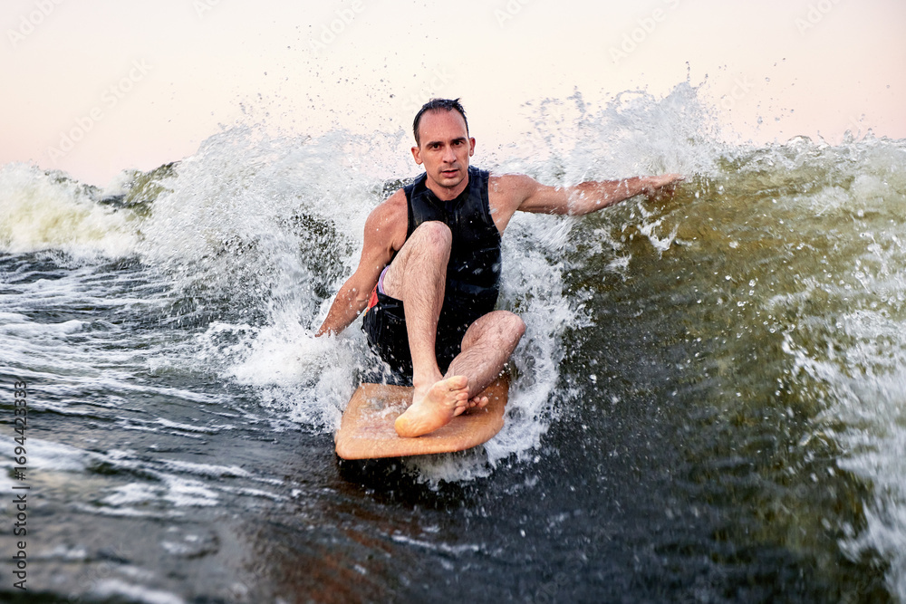 Fototapeta premium Man surfing on wave with wooden board during vibrant sunset