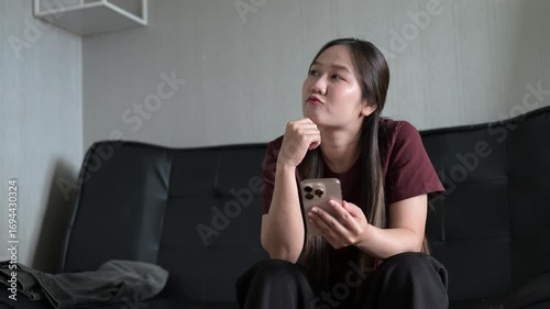 Young Asian woman sitting on sofa using mobile phone while planning and thinking