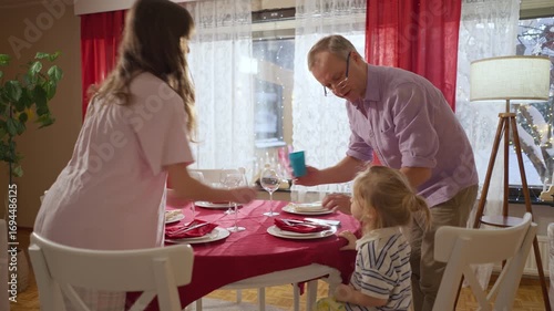 Happy family preparing festive dinner together at home, decorating the table near Christmas tree with glowing lights. Cozy holiday atmosphere, family lifestyle and winter celebration.
