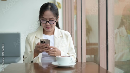 Happy businesswoman sitting in cafe drinking hot mocha from coffee cup smiling looking through window while using phone