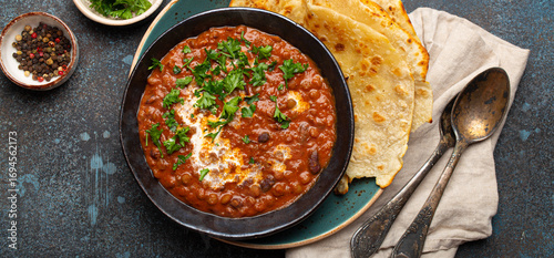 Traditional Indian Punjabi dish Dal makhani with lentils and beans in black bowl served with naan flat bread, fresh cilantro and two spoons on blue concrete rustic table top view.