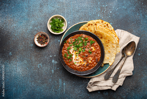 Traditional Indian Punjabi dish Dal makhani with lentils and beans in black bowl served with naan flat bread, fresh cilantro and two spoons on blue concrete rustic table top view. Space for text.