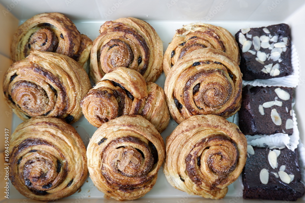 Fototapeta premium Appetizing assortment of freshly baked cinnamon swirl pastries and decadent chocolate brownies, neatly arranged white box. Golden brown spirals and rich fudgy squares offer satisfying dessert