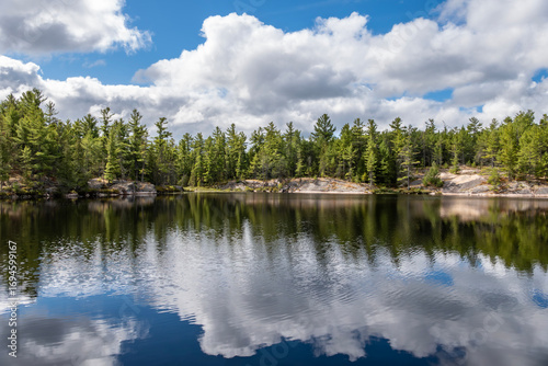Beautiful Reflection of Blue Sky, White Clouds, Evergreen and Pink Granite Rocks on Moira Pond, French River, Ontario