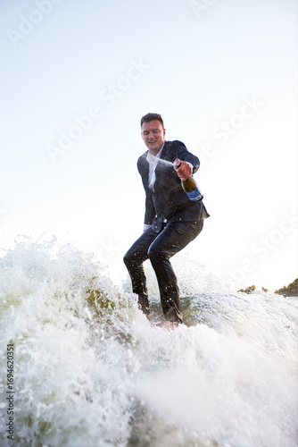 Young man in classic suit with champagne bottle rides a wakeboard on the lake near city. Happy clerk escaped from a stuffy office to take up active sport. Best summer leisure after routine work.