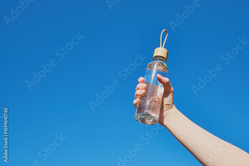 Fotografía Caucasian female hand holding reusable glass bottle of water against blue sky