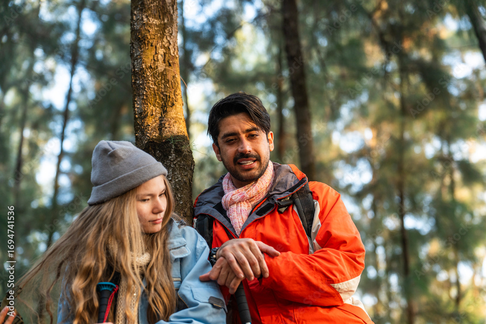 Fototapeta premium Young Couple Resting Against Tree During a Winter Hike. Enjoying the Golden Hour Light in a Peaceful Forest, Wearing Warm Clothes and Backpacks, Sharing a Quiet and Happy Moment Together