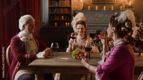 Medium full shot of young 19th century Caucasian gentleman in red frock coat, wig and two mature ladies in fashionable dresses sitting at table in manor parlor, drinking wine with fruits and chatting