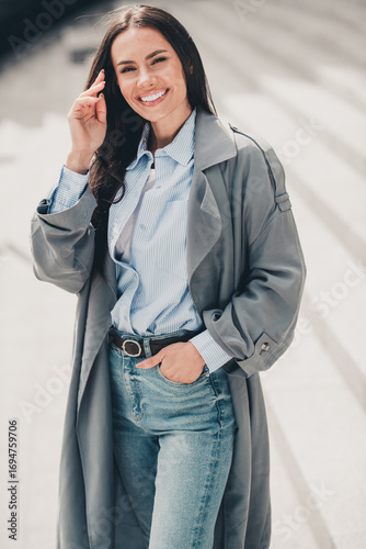 Stylish woman in gray trench coat enjoying an outdoor leisure moment on a city street in a charming urban setting, vertical portrait