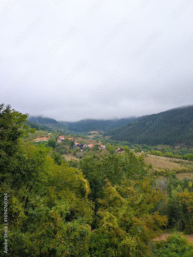 Fototapeta premium Foggy mountains and a valley with small houses and red roofs among green trees. Peaceful rural living and natural beauty in the Balkans.