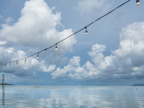 String lights hanging across a calm seaside view with fluffy clouds reflecting on the water under a bright blue sky. Peaceful tropical coastal scenery.