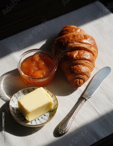 Freshly baked golden croissant with sweet fruit jam and butter on white cloth with natural sunlight illuminating breakfast setting