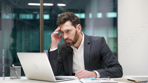 Frustrated businessman having difficulty making transaction using laptop while sitting at workplace in business office. Confused male worker in a formal suit having problems working online on computer