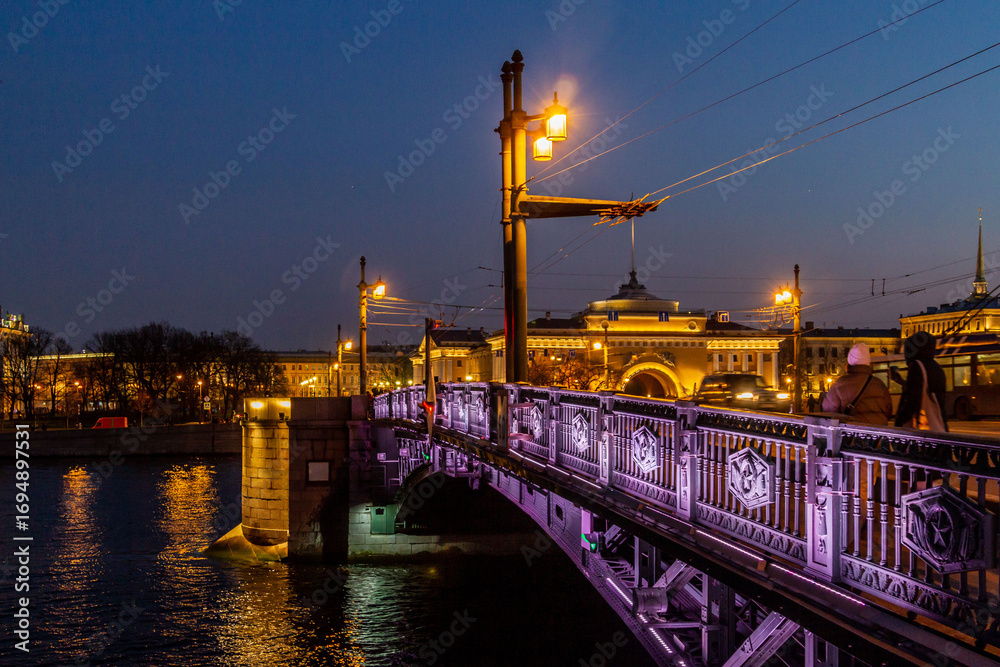 Fototapeta premium Evening view of the Palace Bridge over the Neva River with glowing lanterns, ornate iron railing, and reflections of city lights. Saint Petersburg, Russia.