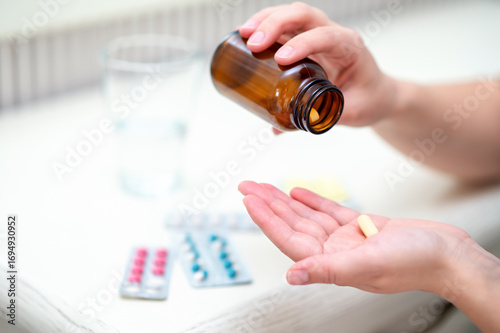 Woman pours pills from amber bottle into her hand with blister packs and a glass of water nearby, concept home medication and daily health routine
