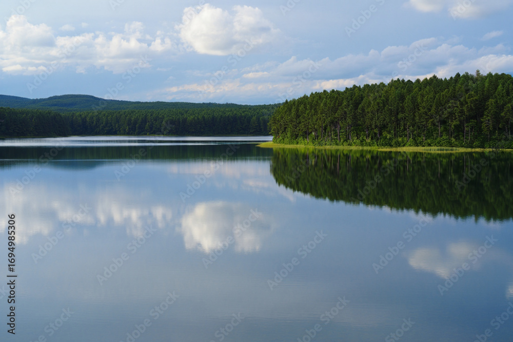 Fototapeta premium Tranquil summer landscape with a forest lake reflecting green trees, a blue sky, and clouds like a mirror