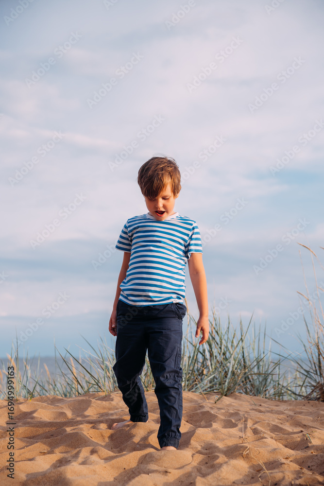 Fototapeta premium Little contented, happy boy on the beach against the background of the sky in the clouds in the grass raising his hands in a sign of success