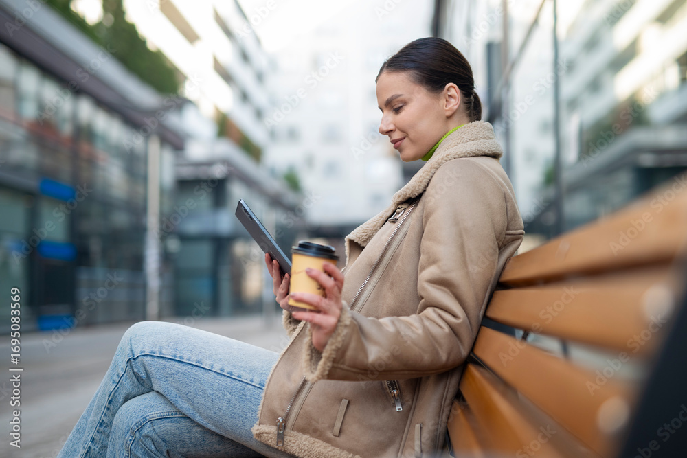 Fototapeta premium Young woman sitting on bench outdoors holding coffee cup and tablet, smiling while reading. 