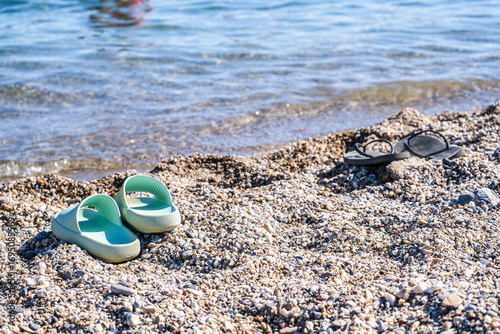 Fototapeta Naklejka Na Ścianę i Meble -  Two pairs of flip flops on a pebble beach with ocean water. Summer holiday shoes by the sea shore in Turkey.