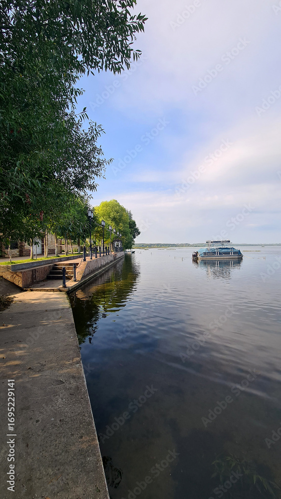 Fototapeta premium A serene lakeside view with a calm water surface reflecting the sky. A boat is anchored near the shore, surrounded by green trees and a concrete walkway.