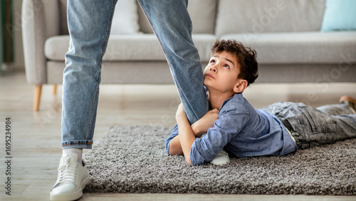 Please stay, dad. Sad little boy embracing his father leg and looking up with begging gaze while lying on the floor carpet. Upset son holding daddy before go to work