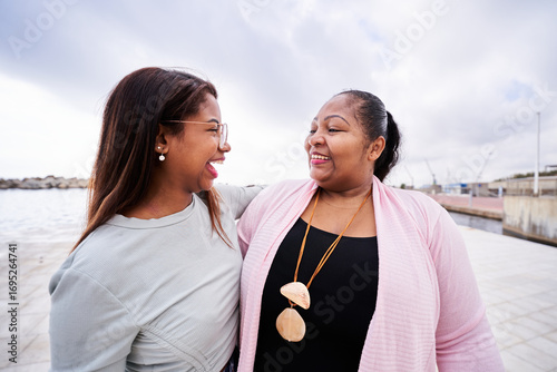 Two cheerful women, possibly mother and daughter, are sharing a joyful moment by the sea, laughing and enjoying each other's company on a cloudy day