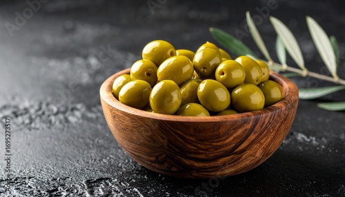 Close-up of a Wooden Bowl Filled with Green Olives and an Olive Branch on a Dark Surface